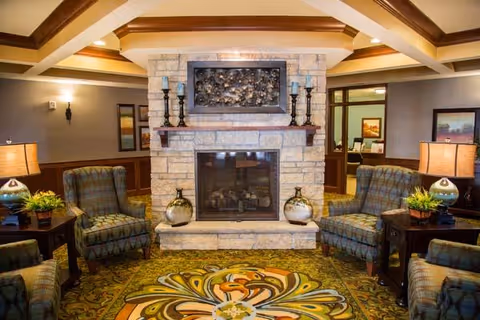 A cozy living room area featuring a stone fireplace with decorative candles and artwork above it. The room has four patterned armchairs arranged around the fireplace, two side tables with lamps and plants, and a colorful patterned carpet. The ceiling has wooden beams and recessed lighting, and there are framed pictures on the walls.