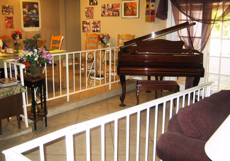 Interior view of a senior living facility showing a polished wooden grand piano with a matching bench near a window with sheer purple curtains. In the background, there is a dining area with wooden chairs and tables decorated with flowers. The room is separated by white metal railings, and there are various pictures and decorations on the walls.