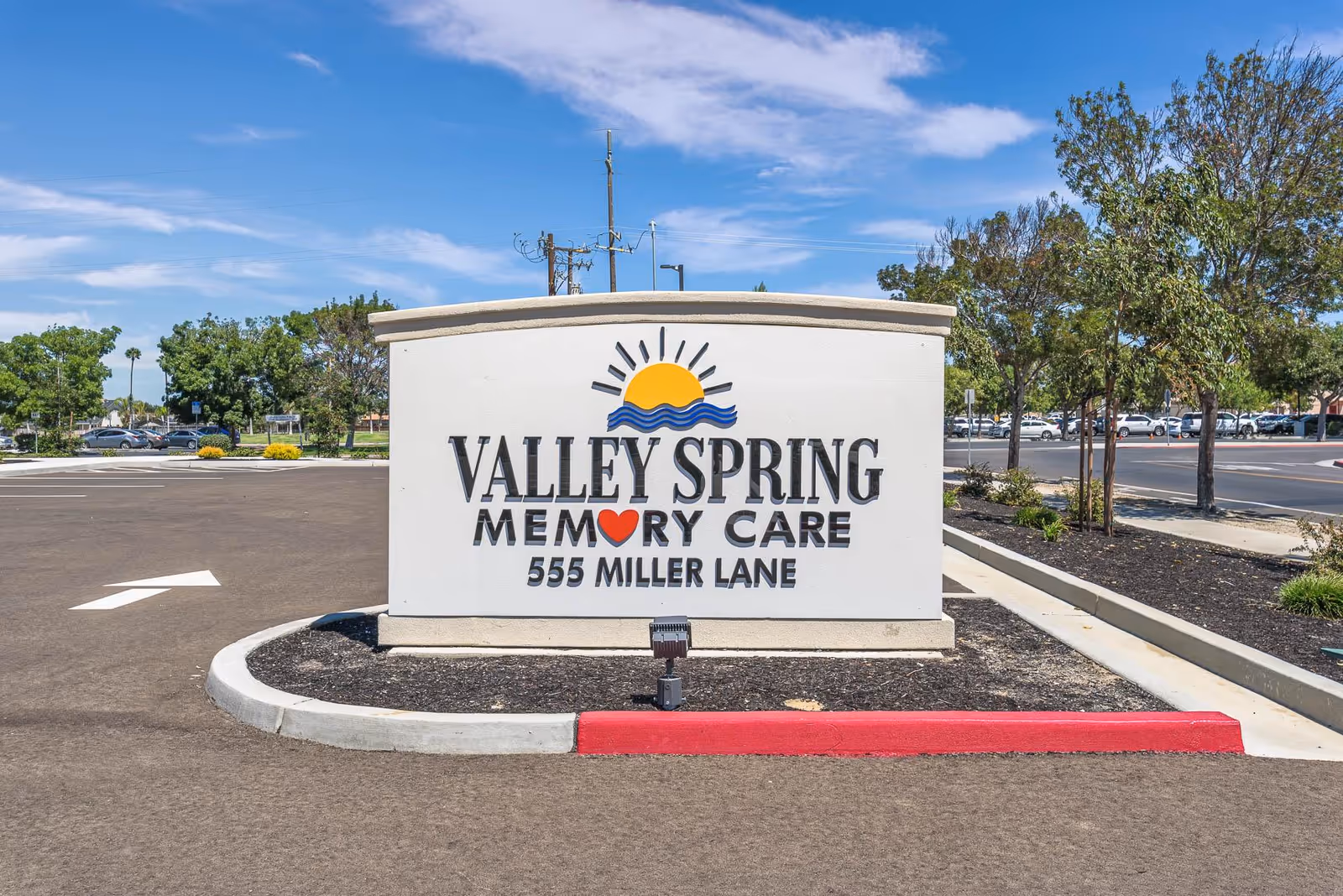 Outdoor view of a large white sign for Valley Spring Memory Care at 555 Miller Lane, with a sun and waves logo above the text. The sign is situated in a landscaped area with trees and a parking lot in the background under a blue sky with some clouds.