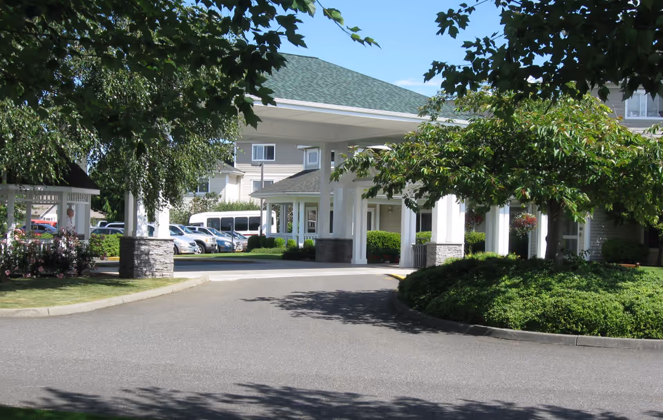 Front entrance and porte-cochere of a senior living facility with a driveway, landscaped shrubbery, and parked vehicles.
