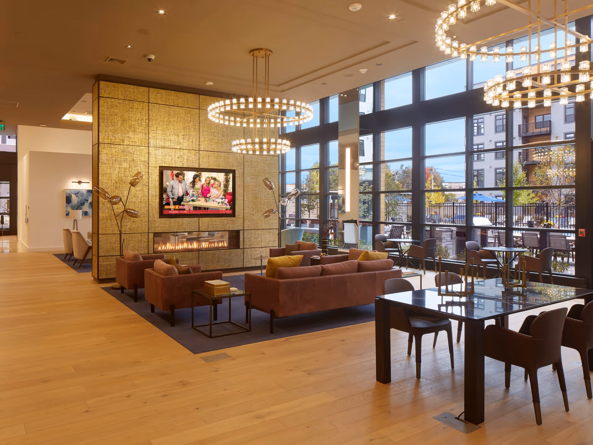 A spacious and modern living room area with large floor-to-ceiling windows letting in natural light. The room features brown sofas and armchairs arranged around a contemporary fireplace with a gold textured wall behind it. Two large circular chandeliers hang from the ceiling. There are also several tables and chairs near the windows, and an outdoor patio area is visible through the glass.