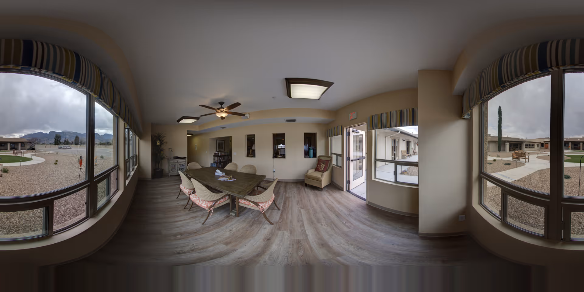 Interior view of a common area in an assisted living facility with large windows showing an outdoor courtyard. The room has a wooden table with six chairs, a ceiling fan with lights, a comfortable armchair near the exit door, and light-colored walls and flooring.