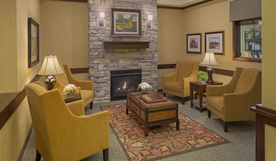A cozy living room with four mustard yellow armchairs arranged around a wooden coffee table on a patterned rug. The room features a stone fireplace with a fire burning, a framed landscape painting above the fireplace, two table lamps on side tables, and framed pictures on the walls.
