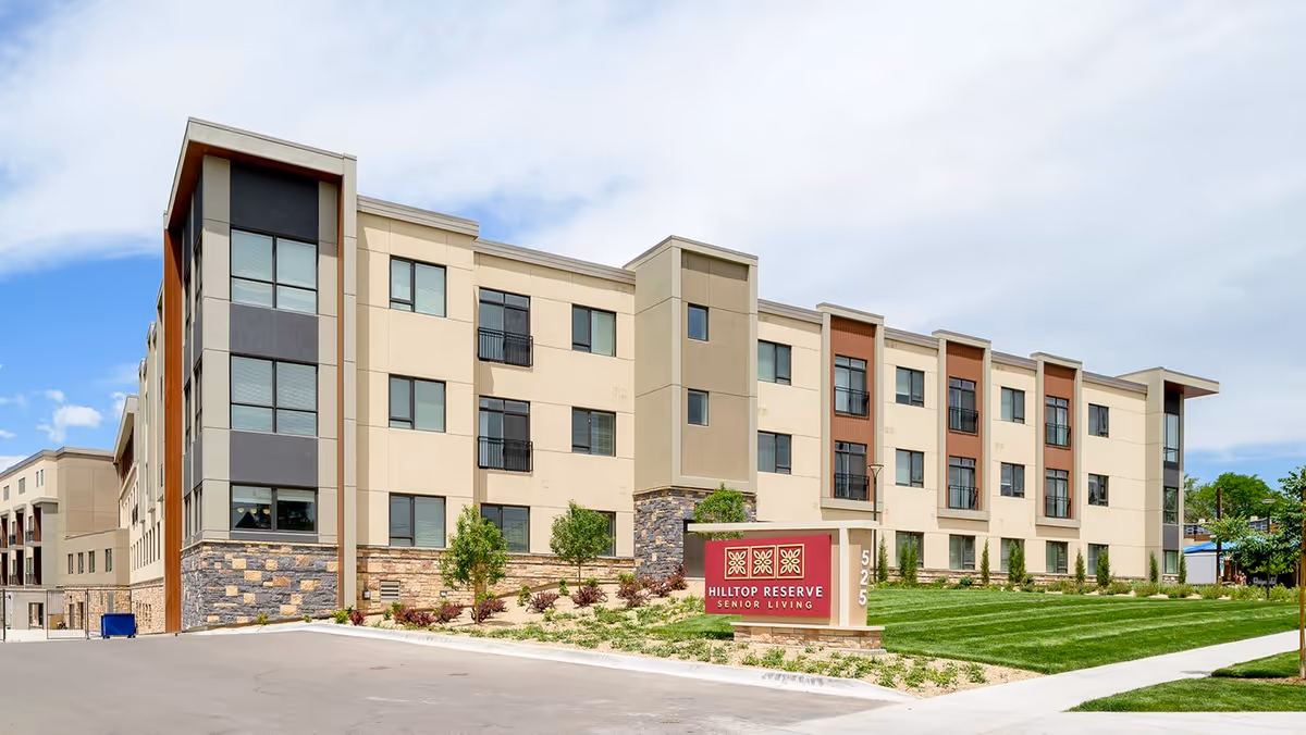 Exterior view of Hilltop Reserve Senior Living, a modern three-story building with beige and gray walls, large windows, and landscaped greenery in front. A red sign with the facility name is visible near the entrance.