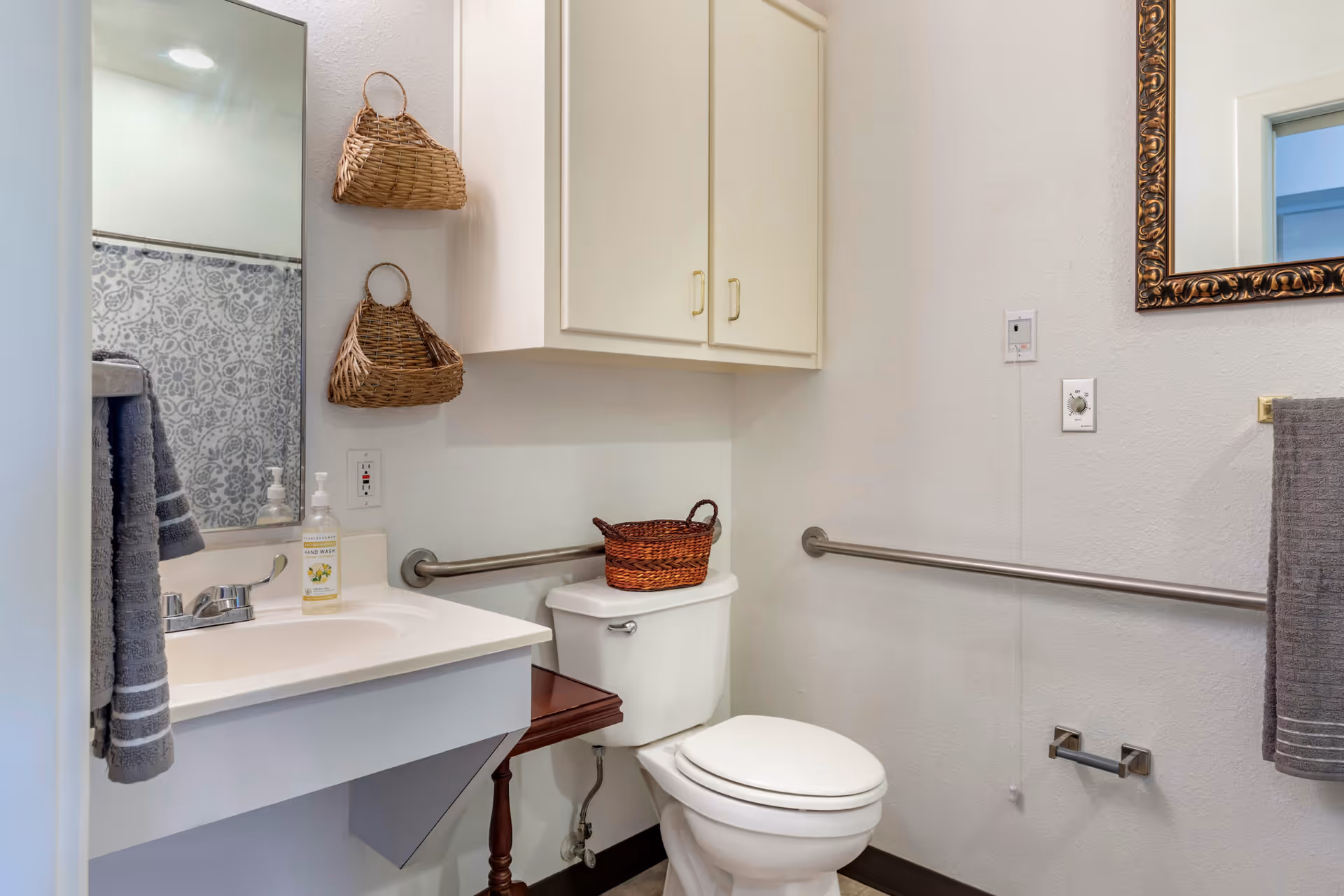 A clean and accessible bathroom featuring a white toilet with a brown wicker basket on top, a white sink with a silver faucet and hand soap, two hanging wicker baskets on the wall, a large mirror above the sink, a towel rack with a gray towel, and grab bars installed on the walls for support.