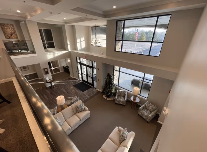 Two-story senior living lobby with sofas and chairs arranged around a Christmas tree, large windows, and the main entrance.