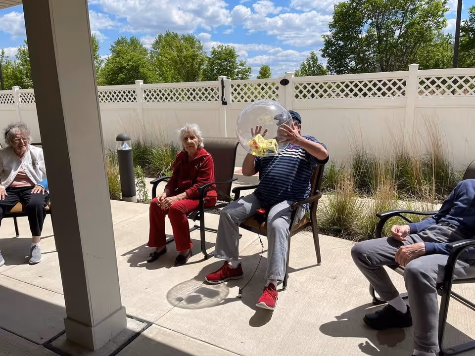 Four elderly individuals sitting on chairs outside on a sunny day near a white fence with greenery in the background. One person is holding a large transparent inflatable ball with yellow cards inside.