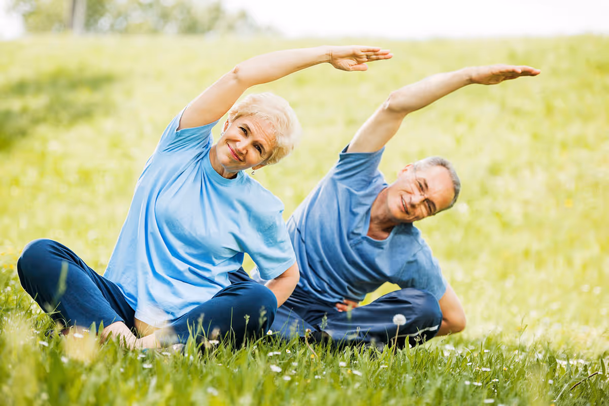 An elderly man and woman sitting on grass outdoors, stretching their arms over their heads while smiling.