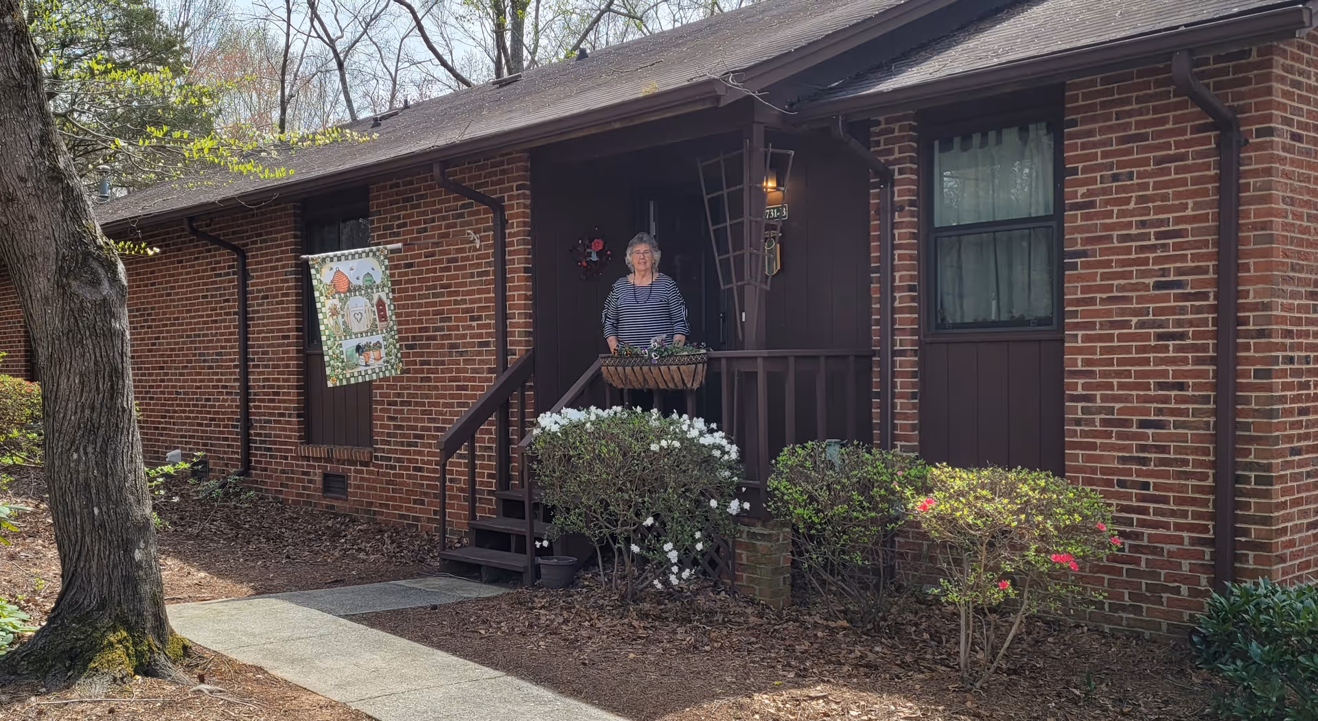 A senior woman standing on the porch of a brick building with dark brown wooden accents. The porch has a small railing and steps leading down to a sidewalk. There are bushes with white and pink flowers in front of the porch, and a decorative flag hanging on the wall. Trees and other greenery surround the building.