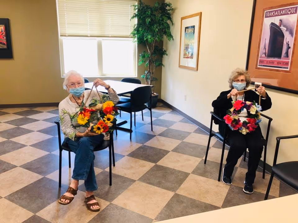 Two elderly women wearing face masks sitting on chairs in a room with checkered flooring, each holding a colorful flower wreath. There is a table with chairs near a window with blinds, a tall potted plant, and framed artwork on the walls.