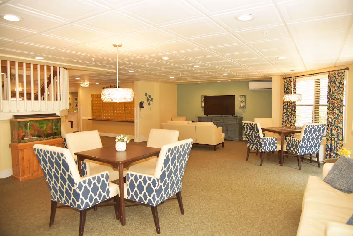 A bright and spacious common area in a senior living facility featuring two square wooden tables each surrounded by four upholstered chairs with blue and white patterned backs. There is a beige sofa in the foreground and another seating area with beige sofas facing a flat-screen TV mounted on a green accent wall. The room has a beige carpet, a white paneled ceiling with recessed lighting, and large windows with floral curtains allowing natural light to enter. A fish tank is positioned near a staircase with white railings, and a wall-mounted phone and mailboxes are visible in the background.