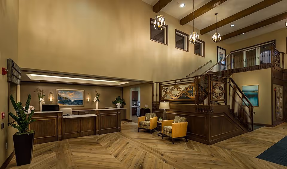 Interior view of a senior living facility lobby with a wooden reception desk, two yellow armchairs with cushions, a wooden staircase with decorative railing, pendant lights hanging from the ceiling, and framed artwork on the walls.