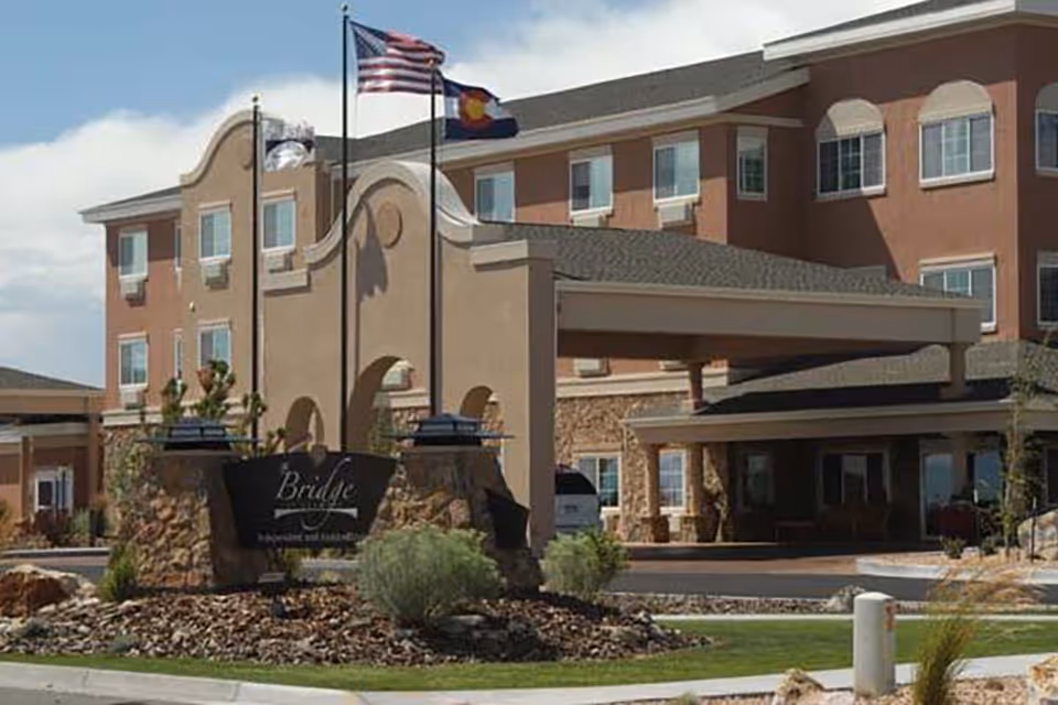 Exterior view of The Bridge at Alamosa senior living facility showing a multi-story building with a covered entrance, stone and stucco facade, landscaping with rocks and shrubs, and three flagpoles with the American flag, Colorado state flag, and another flag.