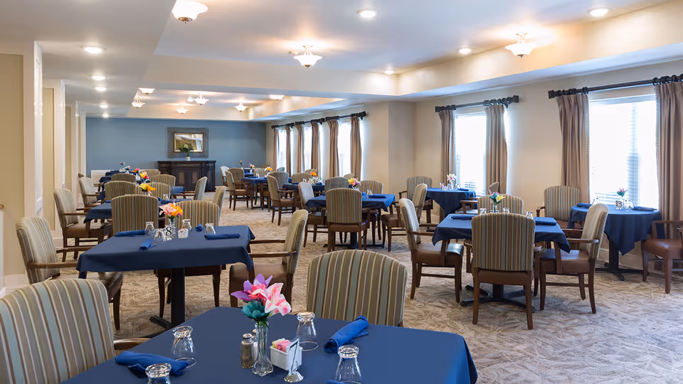 A spacious dining room with multiple tables covered in blue tablecloths, each set with glasses, napkins, and small flower arrangements. The room has beige walls, large windows with brown curtains, and soft overhead lighting. Chairs with striped upholstery surround the tables.