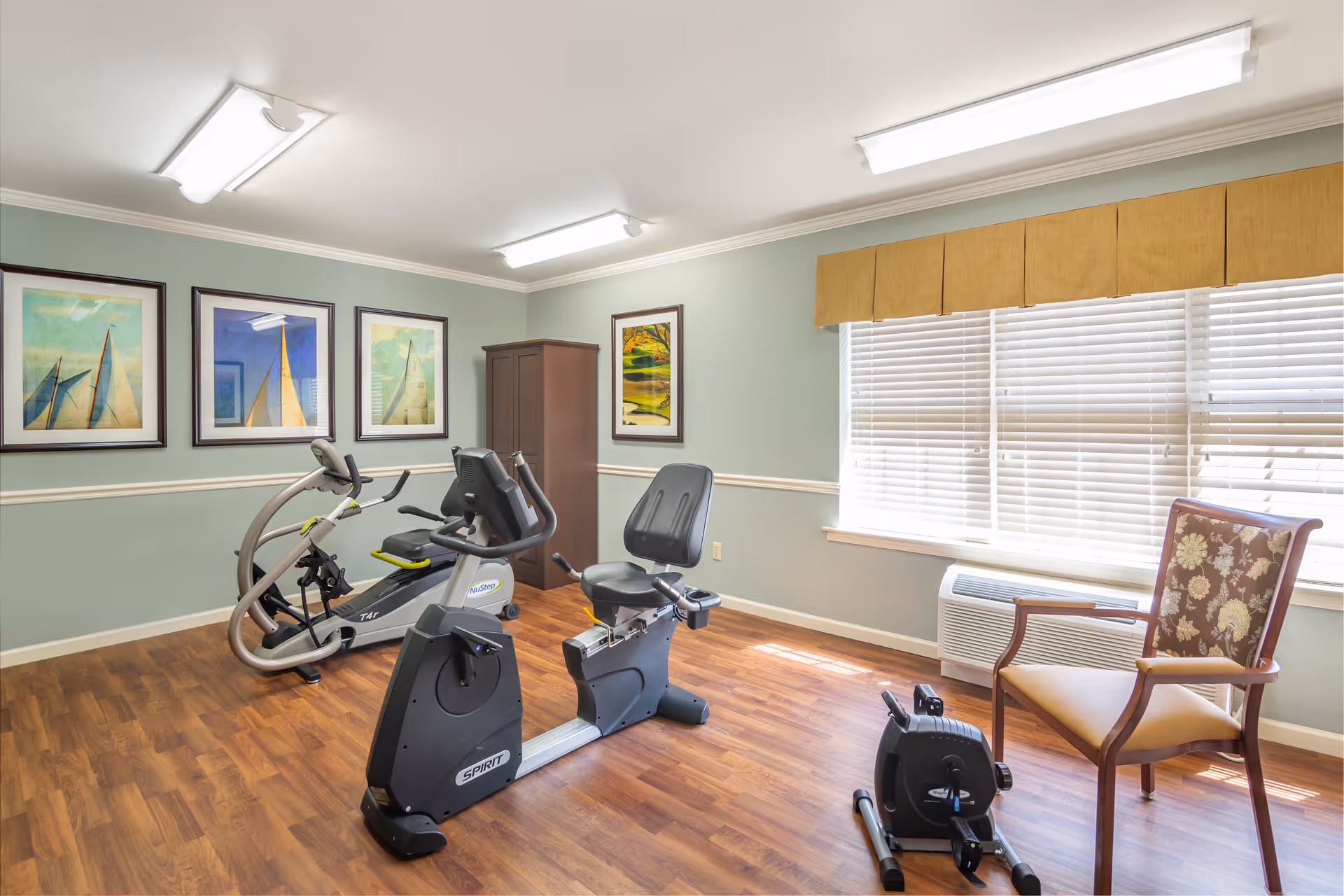 A small exercise room with wooden flooring and light green walls. The room contains two stationary exercise bikes and a small pedal exerciser. There are four framed pictures on the walls, three depicting sailboats and one showing a nature scene. A wooden cabinet is placed in the corner, and a floral-patterned chair is positioned near a window with white blinds and a beige valance. The room is lit by ceiling fluorescent lights.