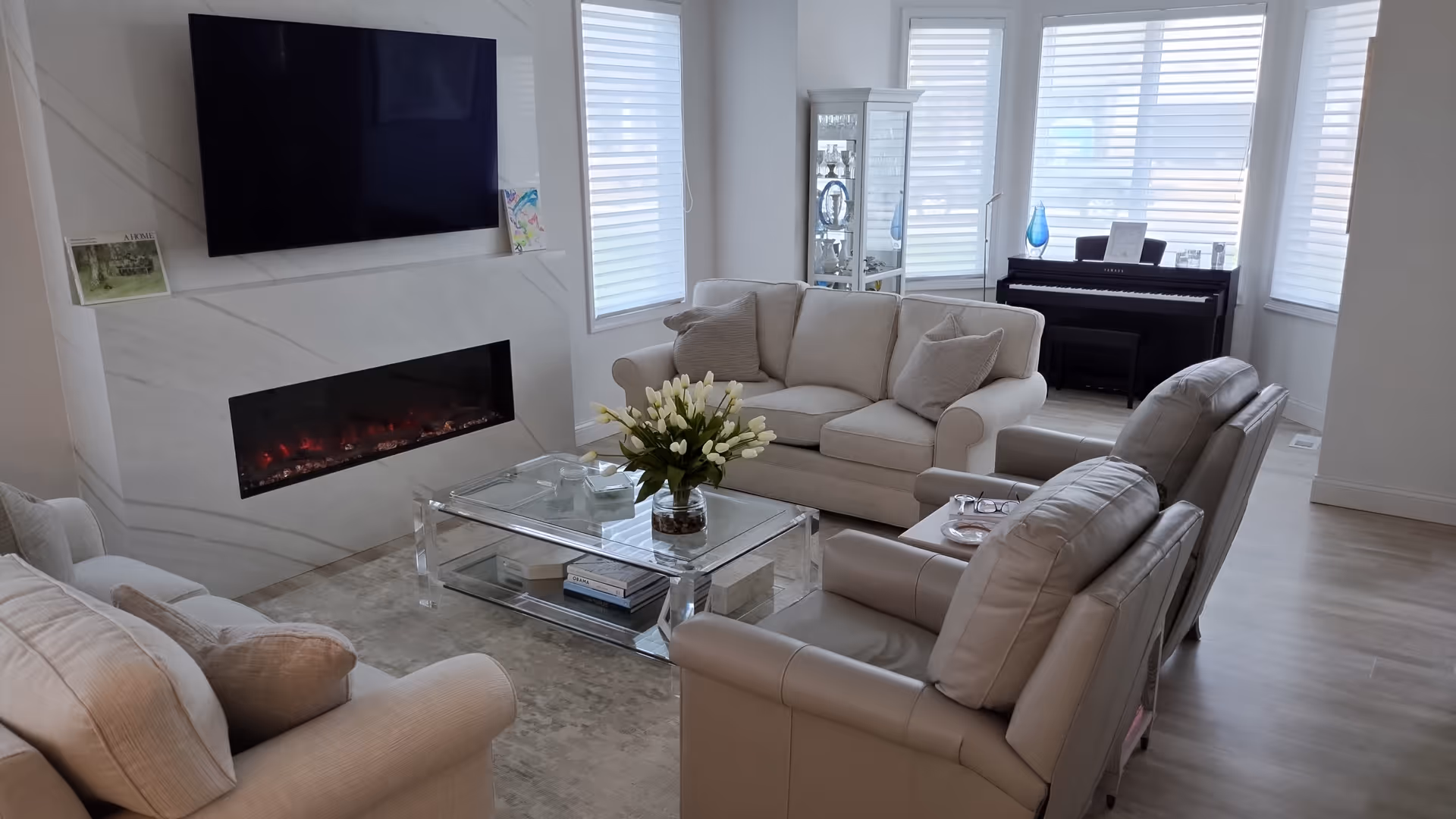 A bright and modern living room with a mounted flat-screen TV above a sleek electric fireplace. The room features a beige sofa, two beige armchairs, a glass coffee table with a vase of white flowers, and a piano near large windows with white blinds.
