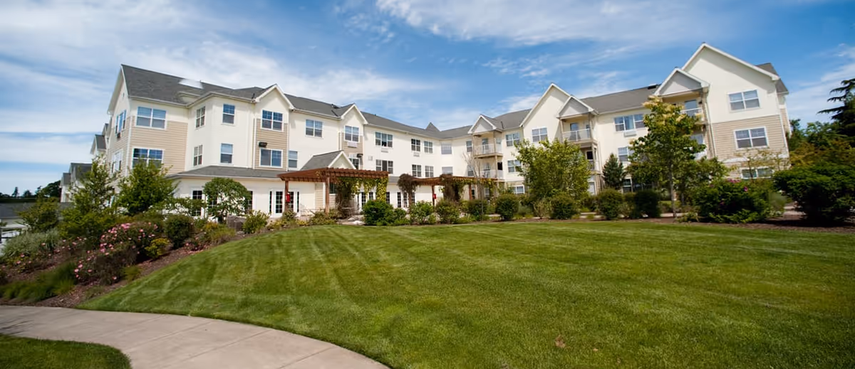 Exterior view of a large, multi-story senior living facility building with beige and white siding, multiple windows, and a well-maintained lawn with shrubs and trees under a partly cloudy blue sky.