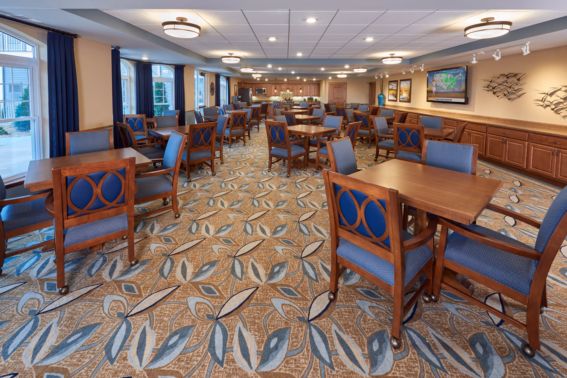 Spacious communal dining room with multiple wooden tables and blue-upholstered chairs, patterned carpet, and windows along one side.