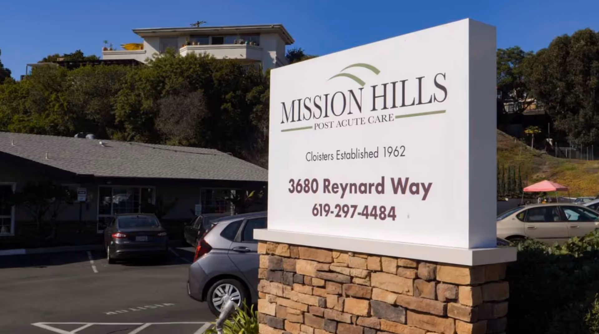 Outdoor view of the entrance sign for Mission Hills Post Acute Care facility, showing the address 3680 Reynard Way and phone number 619-297-4484, with parked cars and buildings in the background under a clear blue sky.