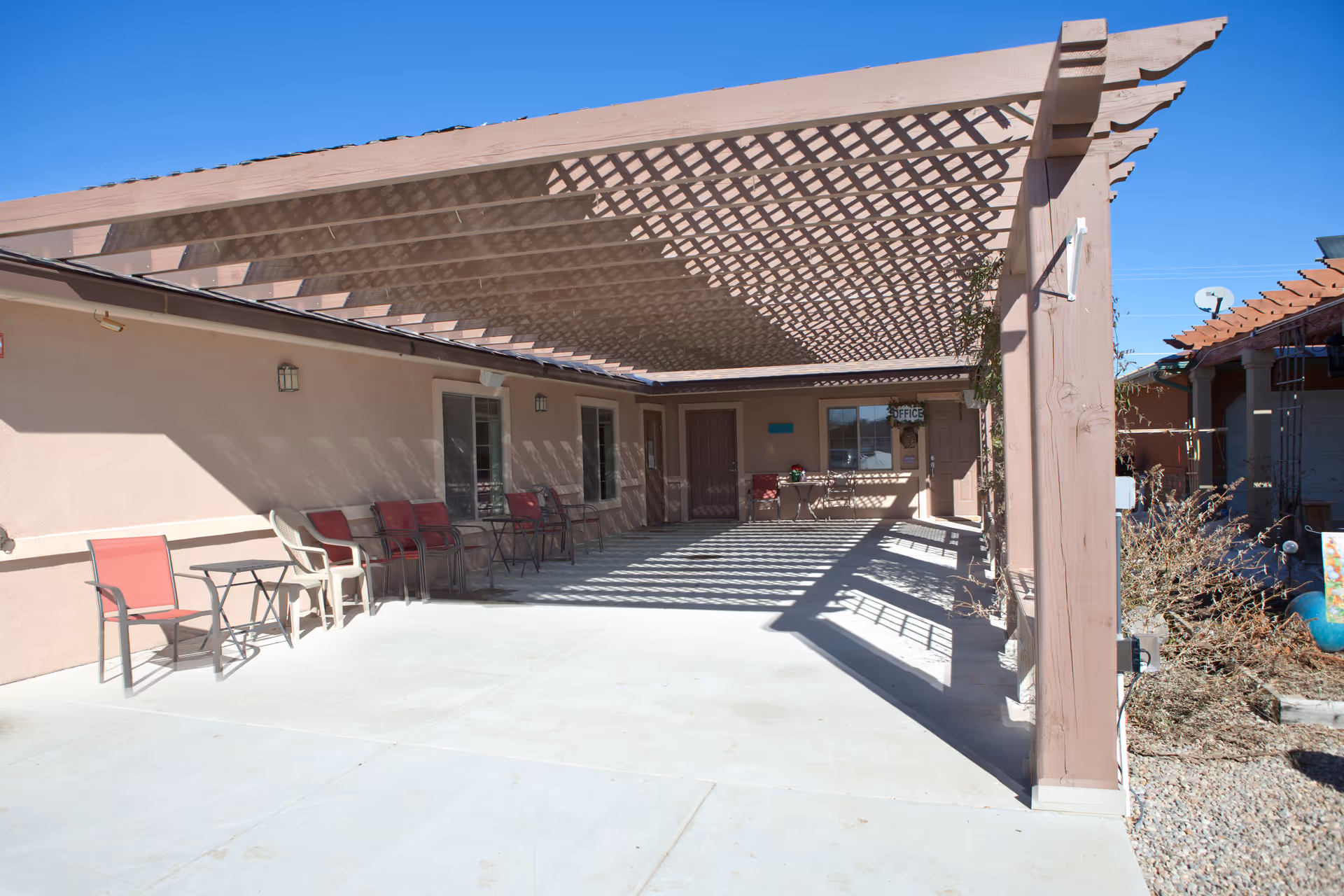Covered outdoor patio with a wooden pergola casting lattice shadows, chairs and tables along the building and an entrance door labeled 'Office'.