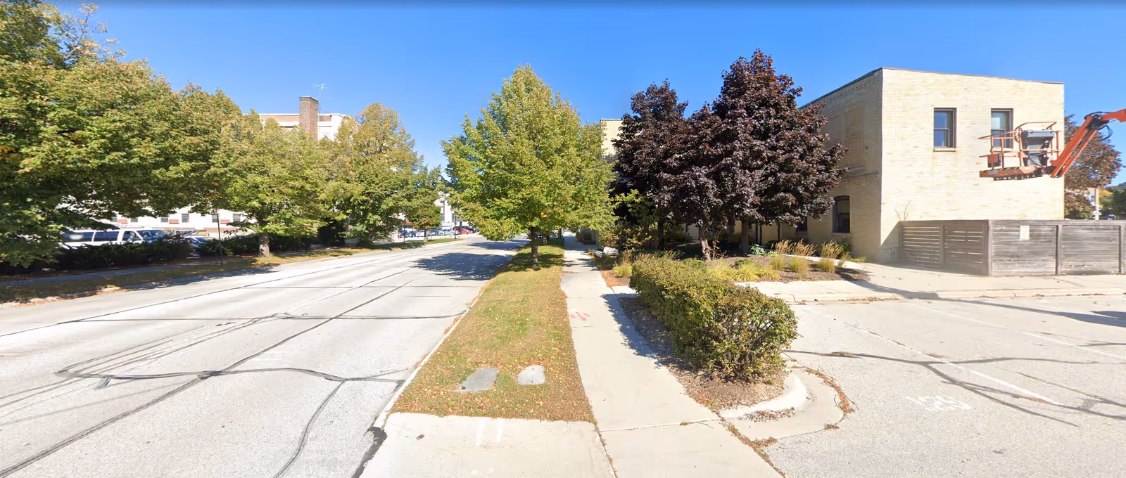 A street view showing a sidewalk lined with trees and bushes on a sunny day. On the right side, there is a light-colored brick building with a construction lift attached to it. The road has visible tire marks and is bordered by grassy areas and more trees.