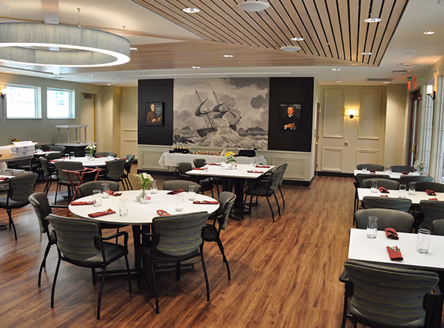 A dining room with multiple round and rectangular tables set with glasses, napkins, and small flower arrangements. The room has wood flooring, a modern ceiling light fixture, and artwork on the walls including a large painting of a ship in a stormy sea.