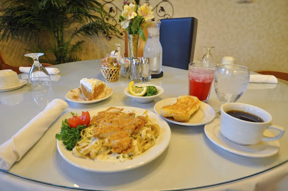 A dining table set with a meal including a plate of pasta with fried chicken, a slice of pie, a small bowl of broccoli with lemon, a piece of toast, a glass of red drink, a glass of water, and a cup of black coffee. The table also has a vase with flowers, salt and pepper shakers, and a basket with condiments. The background shows a decorative wall and a plant.