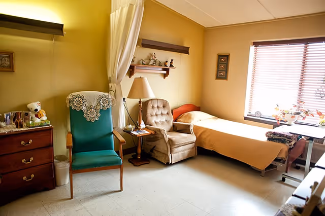 A cozy senior living bedroom with a single bed covered in a beige blanket near a window with blinds. Next to the bed is a beige cushioned armchair and a wooden side table with a lamp and some items. A green upholstered chair with a lace doily on the back is positioned nearby. There is a wooden dresser with drawers and various personal items on top, including a teddy bear. The walls are painted yellow and decorated with small shelves and framed pictures.
