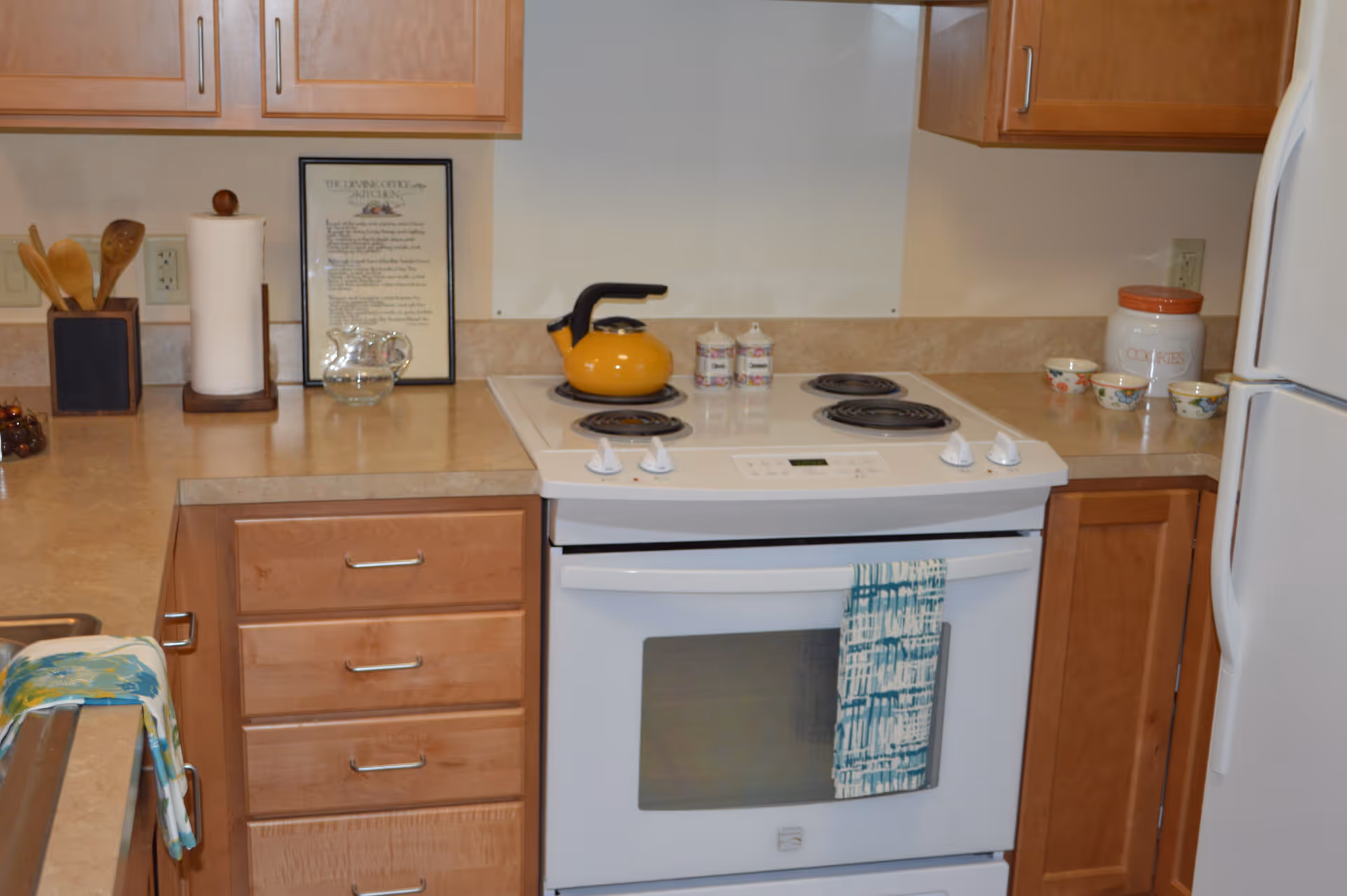 Small kitchen with a white electric stove, wooden cabinets, countertop items and a yellow kettle.
