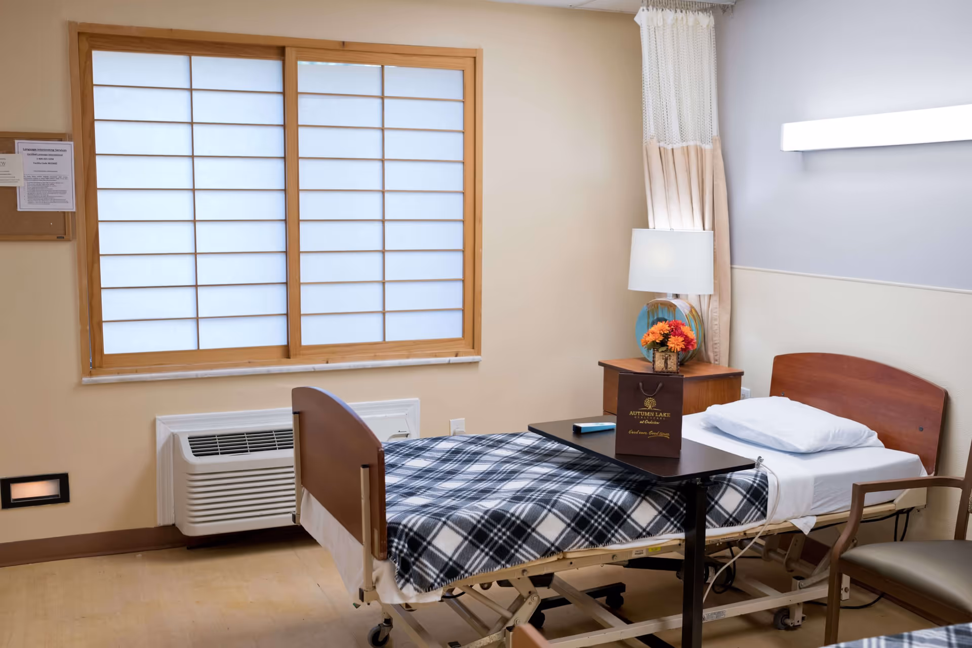 A hospital-style bedroom with a single bed covered with a black and white plaid blanket. There is a small overbed table with a brown Autumn Lake Healthcare at Oakview gift bag and a remote control on it. Next to the bed is a wooden nightstand with a lamp and a small flower arrangement. The room has a window with wooden frame and frosted glass panels, beige walls, and a chair beside the bed.