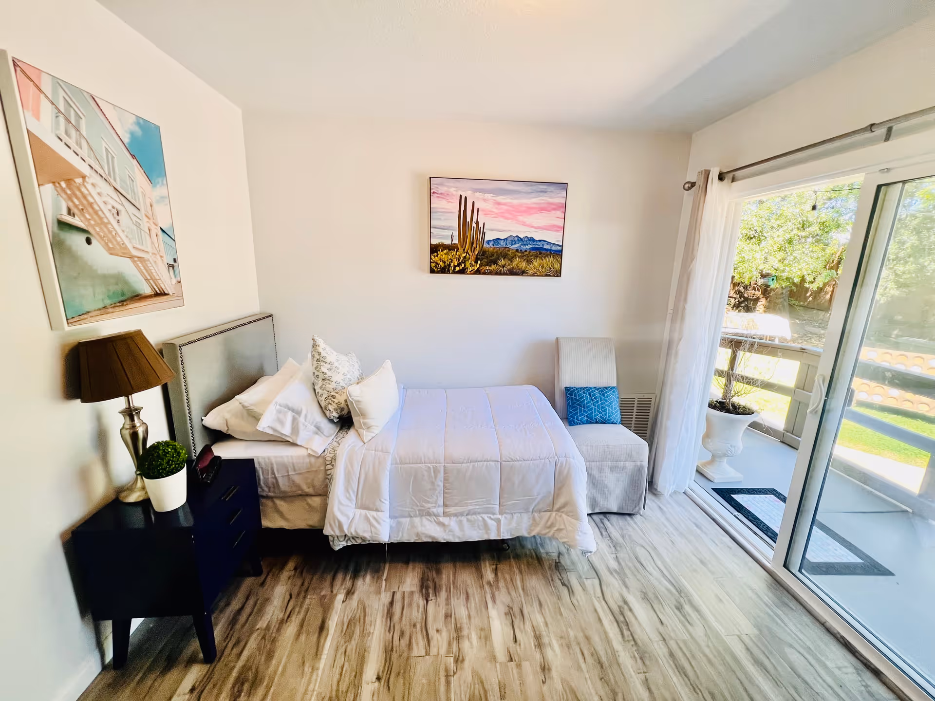 A bright bedroom with a single bed made with white bedding and pillows, a small dark nightstand with a lamp and a potted plant, a cushioned chair with a blue pillow, two framed pictures on the walls, and a sliding glass door leading to an outdoor balcony with greenery visible outside.