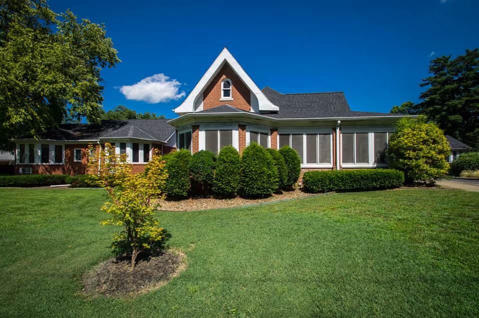 Exterior view of a brick building with multiple windows and a steep triangular roof section, surrounded by green bushes, a small tree, and a well-maintained lawn under a clear blue sky.