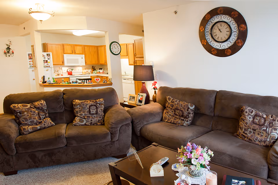 Cozy living room with two brown sofas adorned with patterned cushions, a wooden coffee table with a flower vase, remote control, and decorative items. A large decorative wall clock hangs on the white wall above one sofa. In the background, a kitchen area with wooden cabinets, a microwave, and a refrigerator is visible.