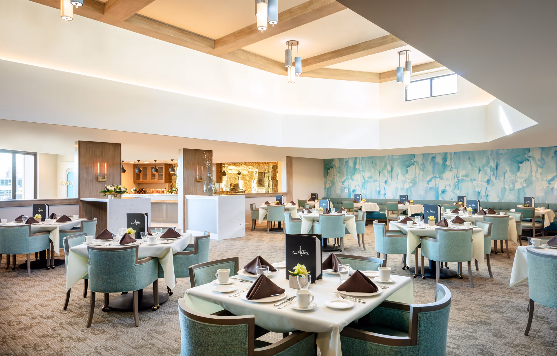 A bright and spacious dining room with multiple round tables set with white tablecloths, brown folded napkins, white cups, plates, and silverware. The chairs are upholstered in a light teal fabric. The room features a high ceiling with wooden beams and modern pendant lights. A large abstract blue and white mural decorates one wall, and there is a serving area with a counter and shelves in the background.