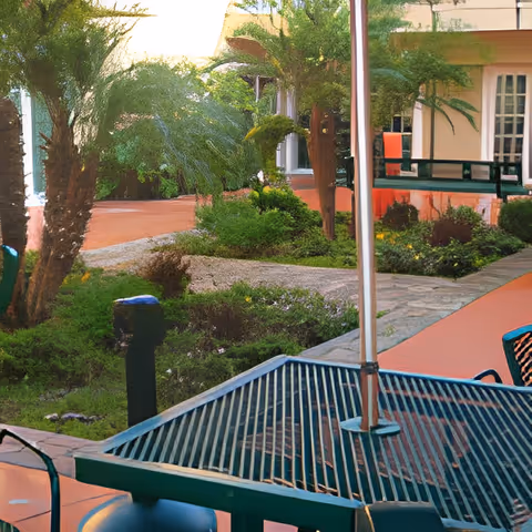 Outdoor patio area with a metal table and chairs, surrounded by greenery including palm trees and bushes, with a building featuring windows and a porch in the background.
