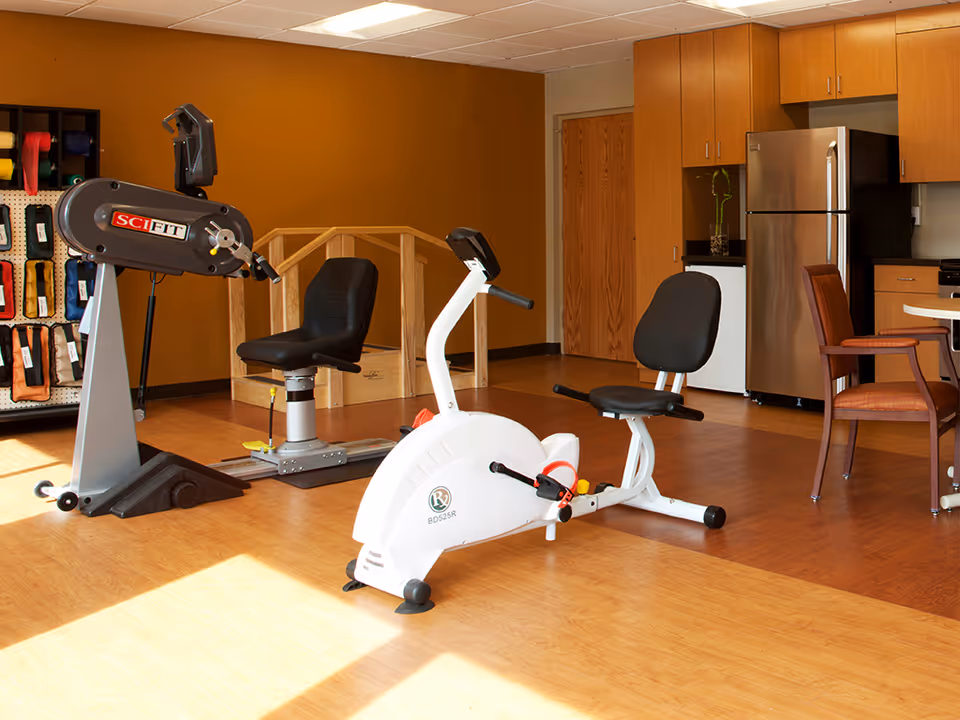 A room with exercise equipment including a recumbent stationary bike and a SciFit machine, wooden flooring, and a kitchenette area with a refrigerator, cabinets, and a table with chairs.