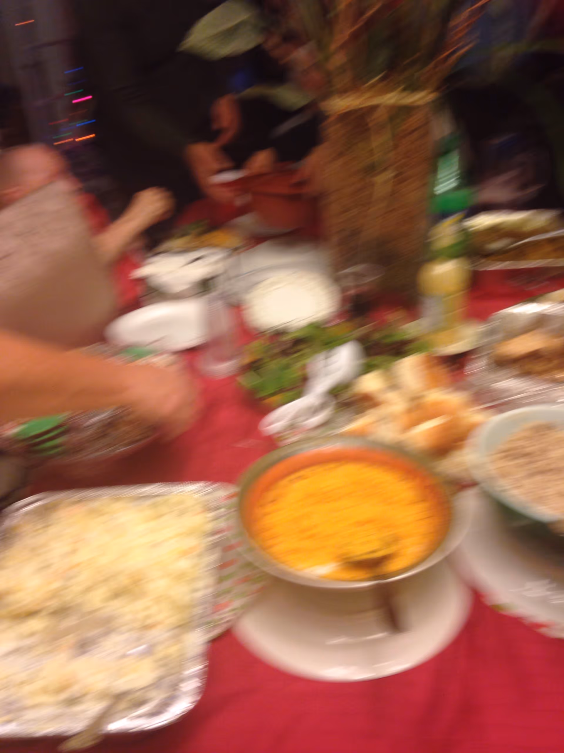 Blurry image of a table covered with a red tablecloth and various dishes of food, including a bowl of orange-colored food, a tray of mashed potatoes, and other plates with food. Several people are partially visible around the table, serving or reaching for food.