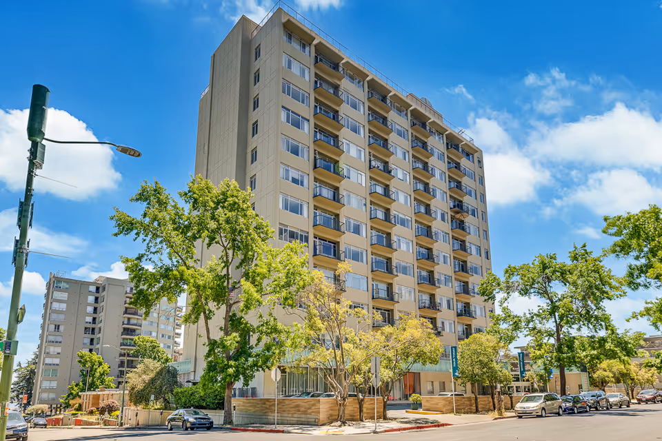 Exterior view of a tall senior living facility building named Lake Park Senior Living on a sunny day with a clear blue sky, surrounded by trees and parked cars along the street.