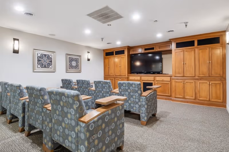 A cozy media room with two rows of patterned armchairs featuring wooden armrests and small attached tables, facing a large flat-screen TV mounted within a wooden built-in cabinet. The room has neutral-colored walls with two framed decorative artworks and modern wall sconces providing soft lighting.