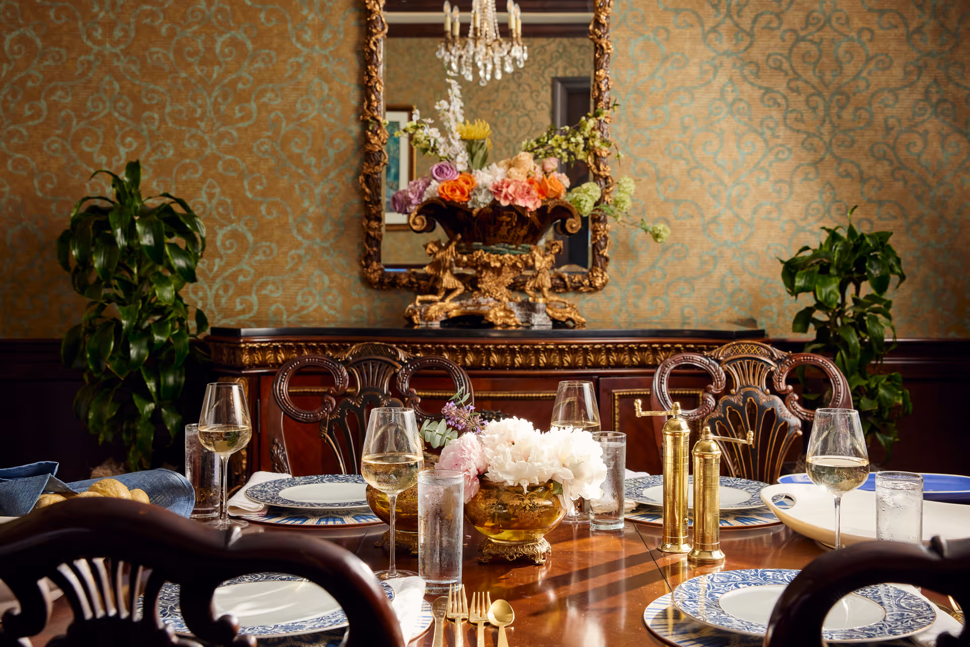 A formal dining room with an ornate wooden dining table set with blue and white patterned plates, gold utensils, wine glasses, and water glasses. A centerpiece with white flowers is on the table. In the background, there is a wooden sideboard with a decorative gold-framed mirror above it and a floral arrangement in a gold vase. Two green plants flank the sideboard, and the walls have a patterned wallpaper.