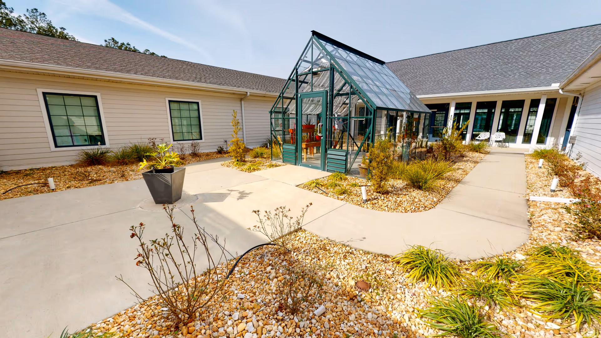 Courtyard featuring a small glass greenhouse at center, concrete walkways, rock landscaping, and surrounding single-story building wings.
