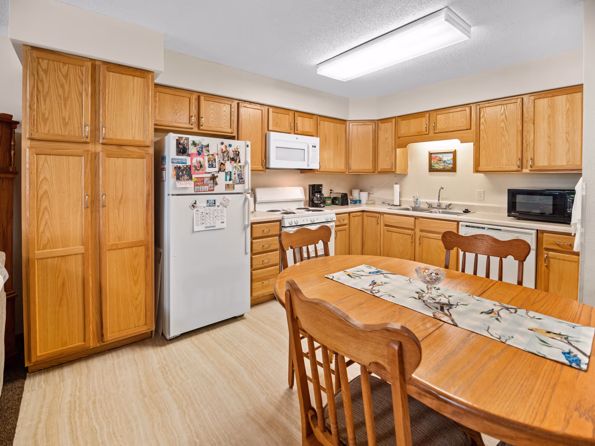 A kitchen with wooden cabinets, a white refrigerator covered with photos and magnets, a white stove with a microwave above it, a coffee maker, a sink, a dishwasher, and a microwave on the counter. In the foreground, there is a wooden dining table with four chairs and a decorative table runner with a bird design.