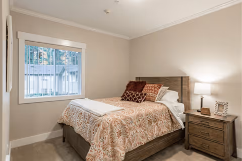 A cozy bedroom with a wooden bed frame and a patterned bedspread in warm tones. The bed is made with multiple pillows and a folded white towel at the foot. Next to the bed is a wooden nightstand with a lamp, a small framed photo, and a clock. A window with vertical blinds shows an outdoor view of trees and a building.