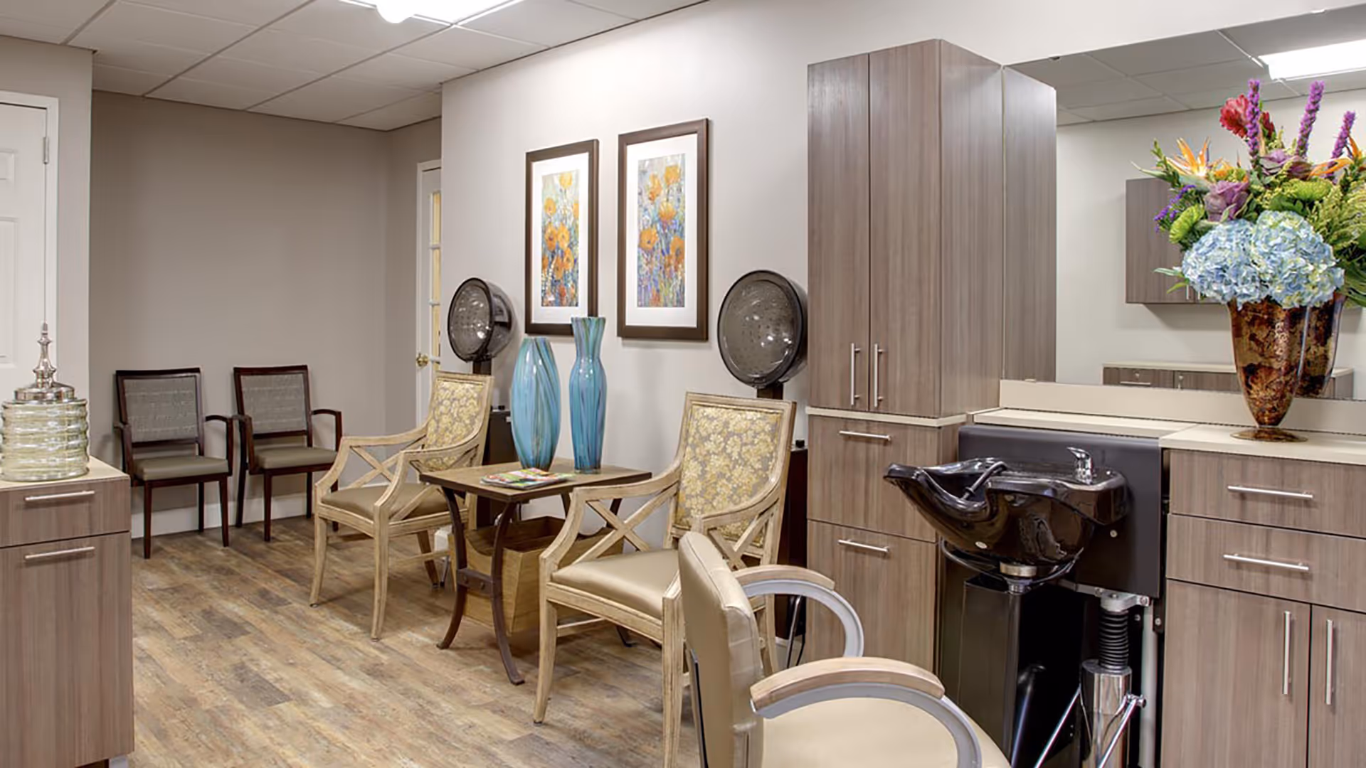 Interior of a salon area in a senior living facility with wooden flooring, two hair drying stations with vintage-style hair dryers, a black hair washing sink, several chairs including two floral upholstered armchairs around a small table with two blue vases, and a large floral arrangement on the counter.