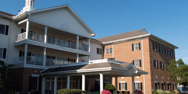 Front entrance of a multi-story senior living building with a covered porte-cochère, balconies, brick and white siding under a clear blue sky.