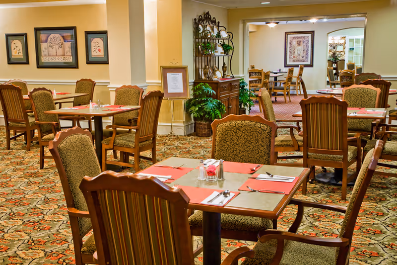 A warmly lit dining room with multiple wooden tables and upholstered chairs arranged neatly. Each table is set with placemats, napkins, silverware, and salt and pepper shakers. The room features patterned carpet, framed artwork on the walls, and decorative plants and furniture along the walls.