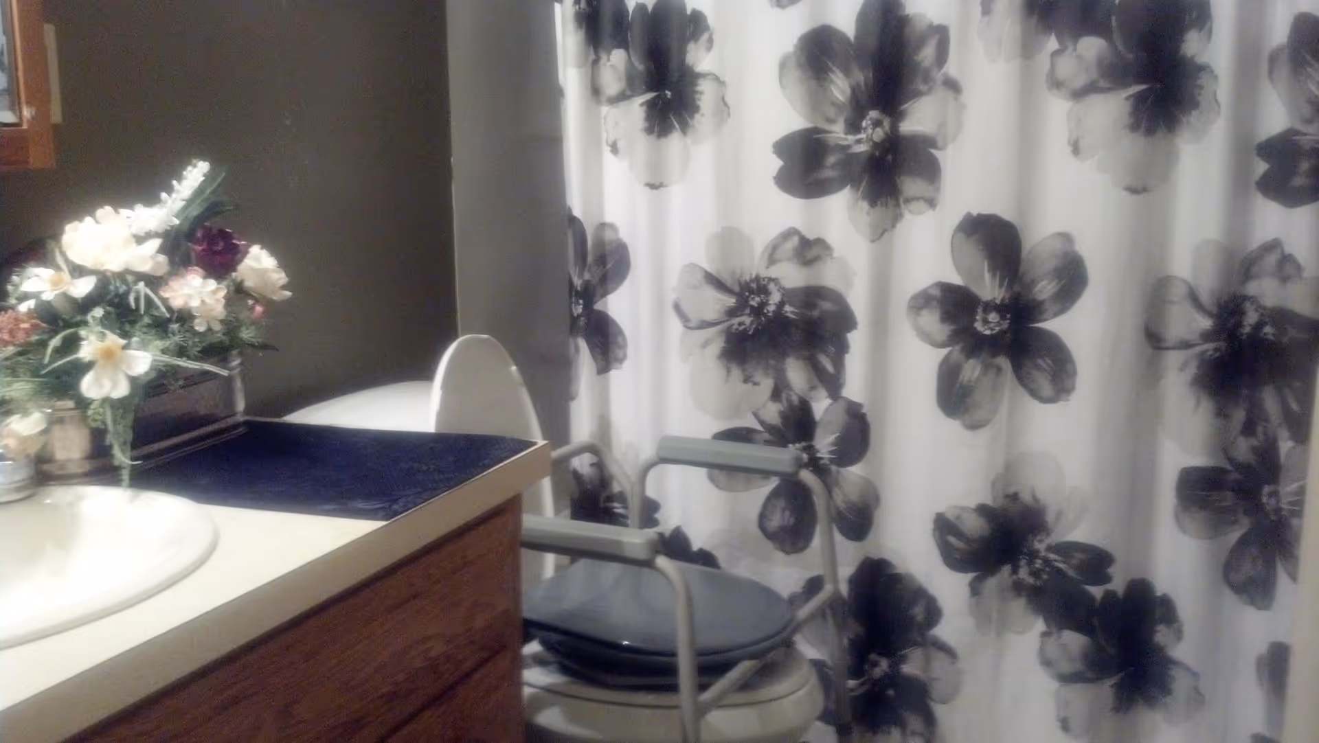 Bathroom with a white sink and wooden cabinet, a floral arrangement on the counter, a toilet with a raised lid, a shower curtain with large black and white flower patterns, and a gray shower chair in front of the shower.