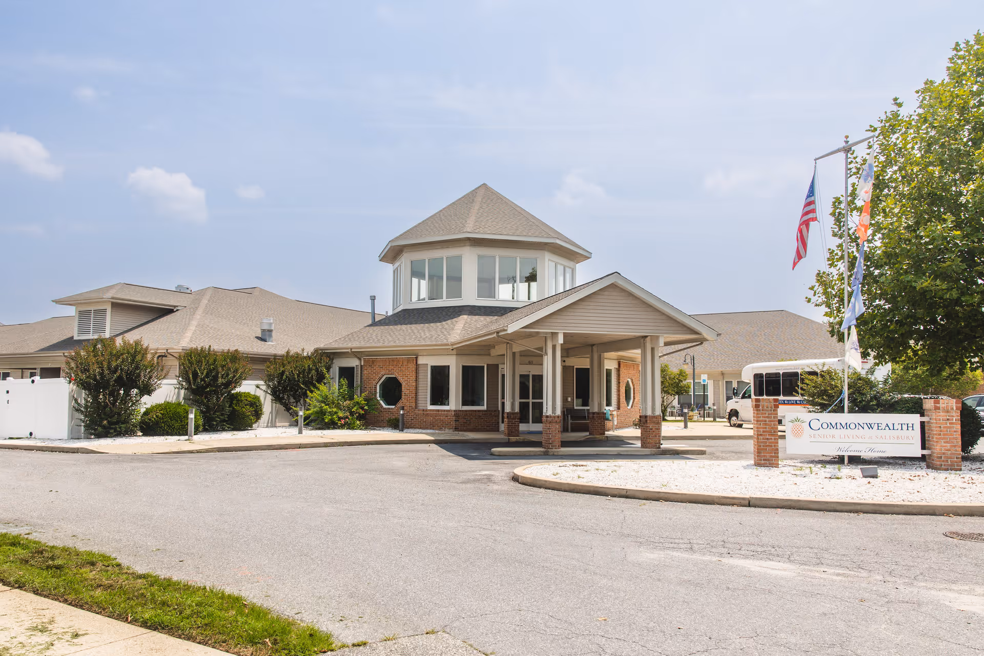 Exterior view of Commonwealth Senior Living At Salisbury building with a covered entrance, brick and beige siding, multiple windows, and a sign with the facility name near the entrance. There are flags on poles and some greenery around the building under a partly cloudy sky.