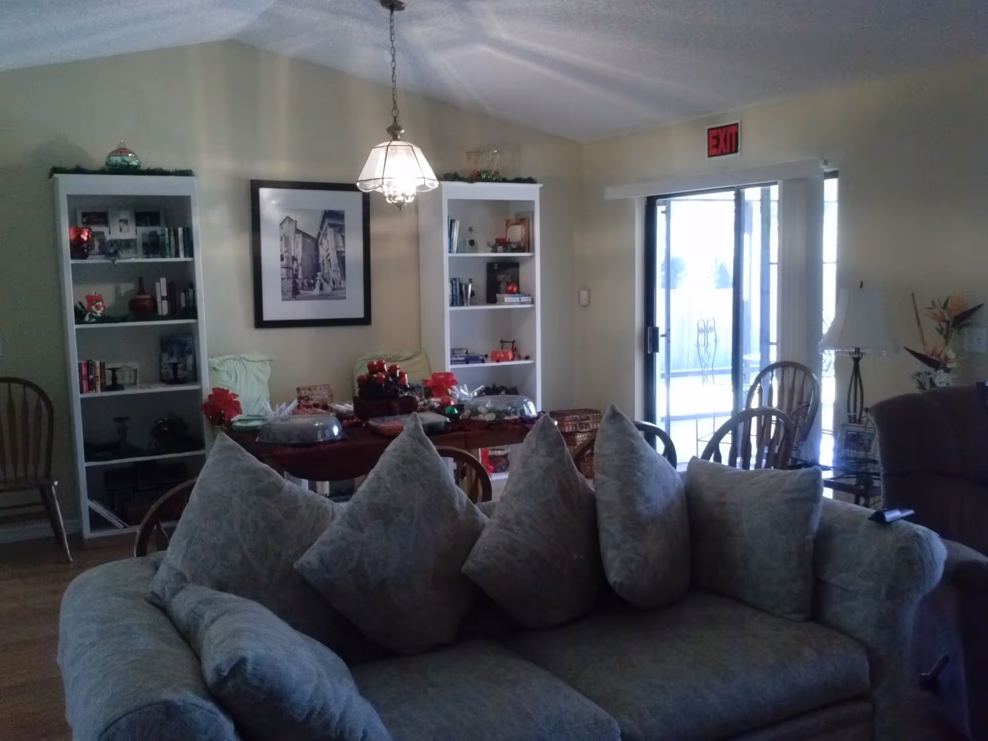 Cozy living room with a large cushioned sofa in the foreground, a dining table and bookshelves against the far wall, and a sliding glass door with an exit sign.