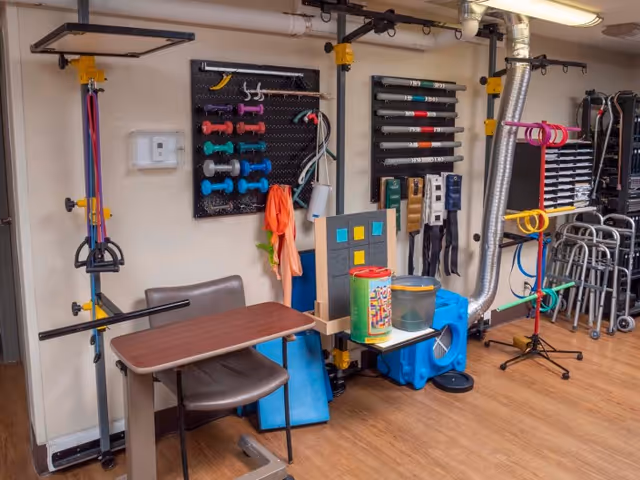 A therapy or rehabilitation room with various exercise equipment including colorful dumbbells, resistance bands, a therapy table with a chair, walkers, and other physical therapy tools organized on the walls and floor.